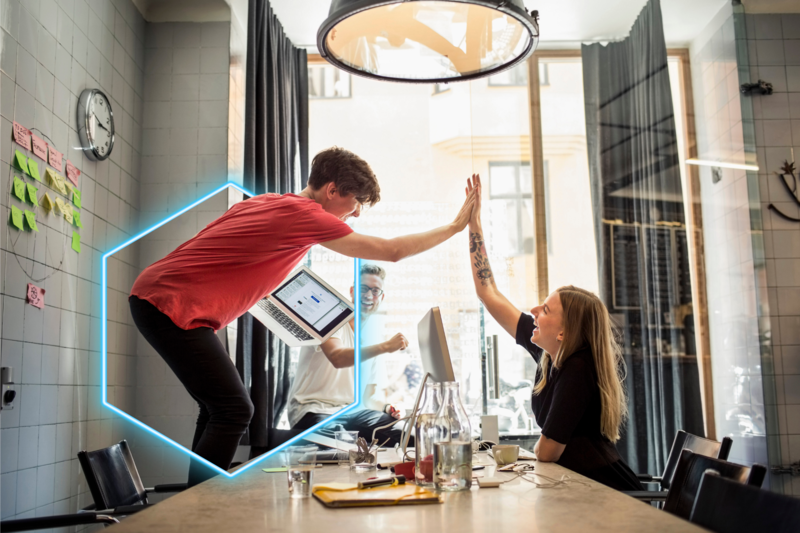 1 persoon met een laptop in zijn hand staat op een stoel en geeft een high-five aan iemand die zit op een stoel. Op de achtergrond kijkt een man lachend toe